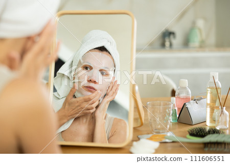Middle-aged woman applying tissue mask on her facial skin sitting in front of the mirror Middle-aged woman applying tissue mask on her facial skin sitting in front of the mirror 111606551