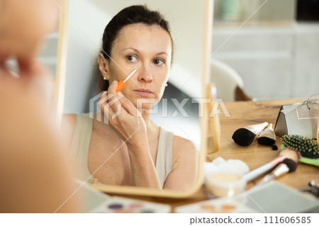 Middle-aged woman applying concealer on her facial skin sitting in front of the mirror Middle-aged woman applying concealer on her facial skin sitting in front of the mirror 111606585