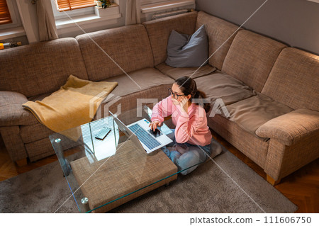 Top view of young Caucasian woman wearing glasses is sitting near sofa and using laptop. Concept of cozy remote work 111606750
