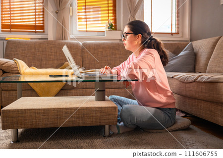 Side view of young Caucasian woman wearing glasses is sitting near sofa and using laptop. Concept of cozy remote work and freelancing Side view of young Caucasian woman wearing glasses is sitting near sofa and using laptop. Concept of cozy remote work and freelancing 111606755