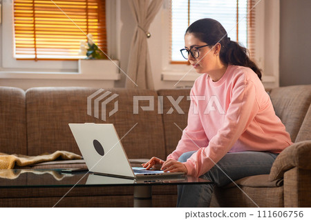 Young Caucasian woman wearing glasses is sitting on couch and using her laptop. Cozy interior. Concept of remote work and freelancing Young Caucasian woman wearing glasses is sitting on couch and using her laptop. Cozy interior. Concept of remote work and freelancing 111606756