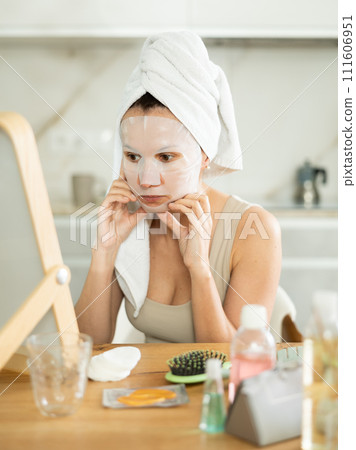 Middle-aged woman applying tissue mask on her facial skin sitting in front of the mirror Middle-aged woman applying tissue mask on her facial skin sitting in front of the mirror 111606951