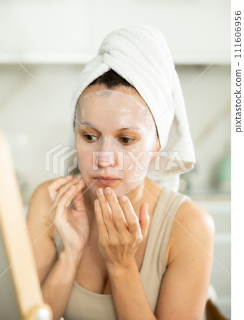 Woman applying moisturizing sheet face mask in front of mirror 111606956