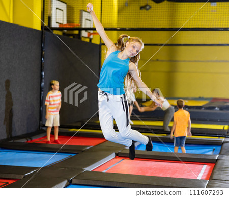 Happy teen girl in blue jersey and white pants jumping and indulging on trampolines in entertainment center 111607293