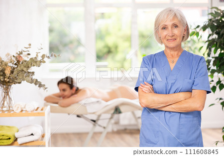 Female elderly physiotherapist poses against background of spa salons office. Female elderly physiotherapist poses against background of spa salons office. 111608845