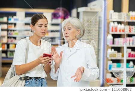 Young girl showing to old pharmacist something displayed on her smartphone Young girl showing to old pharmacist something displayed on her smartphone 111608902