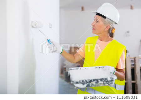 Portrait of elderly positive woman painter in protective helmet and yellow vest who paints walls with paint roller 111609031