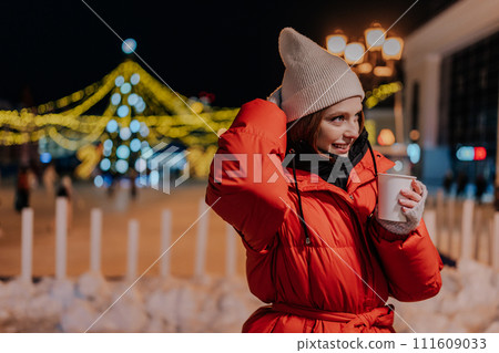 Portrait of smiling young woman wearing warm winter clothes enjoying drinking cup of takeaway coffee outdoors on cold street at night with festive bright illumination. Pretty female walking on park Portrait of smiling young woman wearing warm winter clothes enjoying drinking cup of takeaway coffee outdoors on cold street at night with festive bright illumination. Pretty female walking on park 111609033