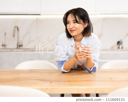 Relaxed Asian girl in male shirt enjoying morning coffee in kitchen Relaxed Asian girl in male shirt enjoying morning coffee in kitchen 111609131