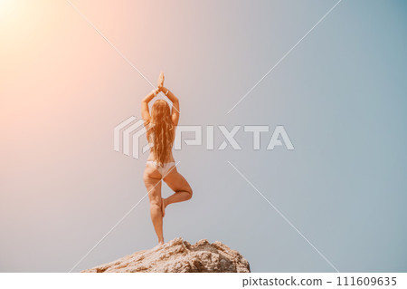 Woman sea yoga. Back view of free calm bliss satisfied woman with long hair standing on top rock with yoga position against of sky by the sea. Healthy lifestyle outdoors in nature, fitness concept. Woman sea yoga. Back view of free calm bliss satisfied woman with long hair standing on top rock with yoga position against of sky by the sea. Healthy lifestyle outdoors in nature, fitness concept. 111609635