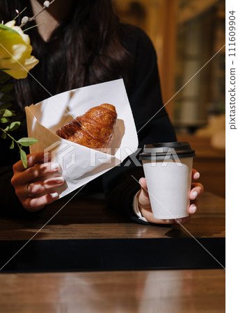 Woman holding a mug of coffee and a croissant in her hands in coffee shop 111609904