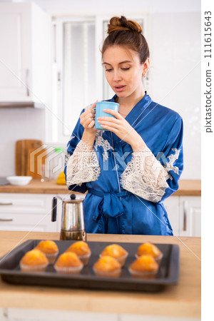 Woman in blue robe drinking coffee near table with cupcakes in home kitchen Woman in blue robe drinking coffee near table with cupcakes in home kitchen 111615643