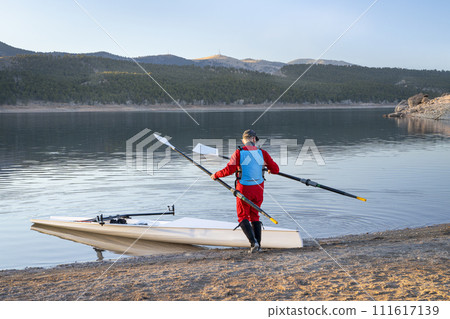 Senior male rower with a coastal rowing shell and hatchet oars on a shore of Carter Lake in fall or winter scenery in northern Colorado. 111617139
