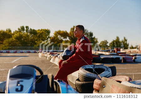 Male racer in protective overalls holding helmet on go-kart track outdoors 111618312