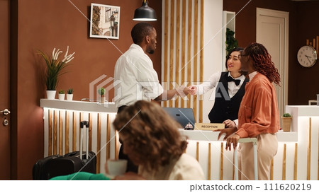 Happy guests on business trip and professional resort employee chatting in cozy stylish hotel lobby. Friendly receptionist handing company coworkers room access key card during check in process. 111620219