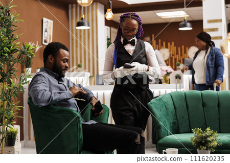African American man tourist sitting in hotel lobby paying for coffee with smartphone, making contactless payment. Friendly waitress holding pos terminal serving guest at luxury resort lounge area 111620235