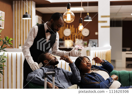 Hotel concierge trying to wake up sleeping tourists in lobby inviting guests to check-in. Exhausted tourists taking rest while waiting for room, feeling sleepy after long-distance travel 111620306