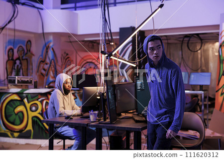 Suspicious hacker looking away after breaking into server and doing illegal activity. Young asian criminals in hood leaving abandoned warehouse after breaching database and stealing data 111620312