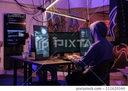 Hacker in hood using spyware to steal information from server while coding on computer with multiple screens. Man programming illegal malicious software in hideout place Hacker in hood using spyware to steal information from server while coding on computer with multiple screens. Man programming illegal malicious software in hideout place 111620340