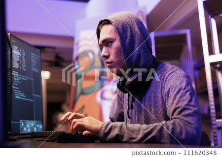 Asian criminal breaking into database and stealing confidential data. Hacker in hood using internet virus malware on computer to breach data while working in abandoned warehouse at night Asian criminal breaking into database and stealing confidential data. Hacker in hood using internet virus malware on computer to breach data while working in abandoned warehouse at night 111620348