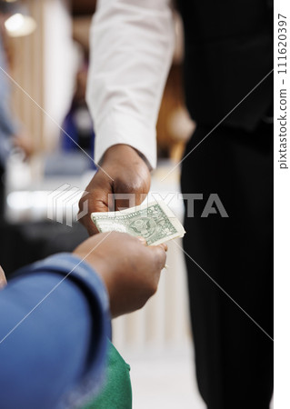 Close up of male African American waiter receiving tip from generous hotel guest. Tourist giving money cash to restaurant worker, making order, paying bill. Tipping in hospitality industry Close up of male African American waiter receiving tip from generous hotel guest. Tourist giving money cash to restaurant worker, making order, paying bill. Tipping in hospitality industry 111620397