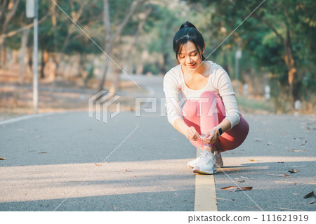 Fitness and running concept, Active young woman pauses to tie her shoelace on a sunlit park path during a workout. Fitness and running concept, Active young woman pauses to tie her shoelace on a sunlit park path during a workout. 111621919