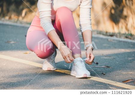Fitness and running concept, A detailed view of a woman in pink leggings tying her shoe on a paved park path, focusing on health and fitness. Fitness and running concept, A detailed view of a woman in pink leggings tying her shoe on a paved park path, focusing on health and fitness. 111621920