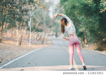 Fitness and running concept, Back view of a woman stretching arms upwards, preparing for a run on a tranquil park road surrounded by trees. Fitness and running concept, Back view of a woman stretching arms upwards, preparing for a run on a tranquil park road surrounded by trees. 111621921