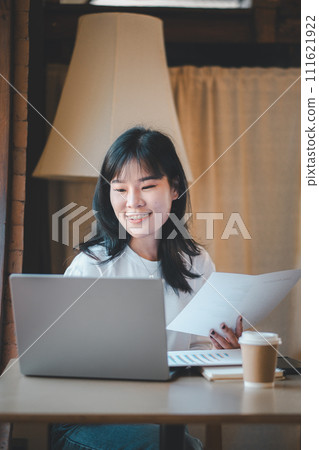 Business freelance concept, A content young Asian woman reviews paperwork with a smile, seated at a cafe with her laptop and a cup of coffee. Business freelance concept, A content young Asian woman reviews paperwork with a smile, seated at a cafe with her laptop and a cup of coffee. 111621922