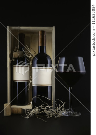 Two red wine bottles with mock up in a wooden box next to a full wine glass and decorative straw, on a dark background. Two red wine bottles with mock up in a wooden box next to a full wine glass and decorative straw, on a dark background. 111623084