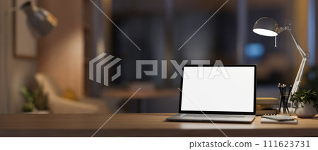 A modern, cozy home office at night with a white-screen laptop computer mockup on the table. 111623731