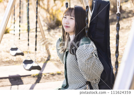 A woman carrying a guitar sitting on a swing in the park and looking at the scenery 111624359