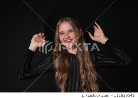 Portrait happy young woman with toothy smile looking at camera and holding brim of felt hat with both hands. Woman poses playfully on black background. Model with long hair dressed in black knit dress Portrait happy young woman with toothy smile looking at camera and holding brim of felt hat with both hands. Woman poses playfully on black background. Model with long hair dressed in black knit dress 111629541