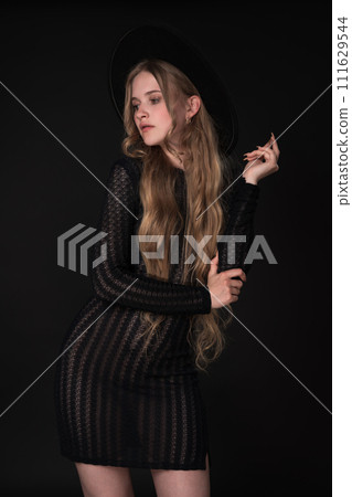 Portrait of shy young blonde looking away, shyly raising one arm bent at elbow, supporting it with other. Blonde with long hair wears short black tight knitted see-through dress, wide brimmed felt hat Portrait of shy young blonde looking away, shyly raising one arm bent at elbow, supporting it with other. Blonde with long hair wears short black tight knitted see-through dress, wide brimmed felt hat 111629544