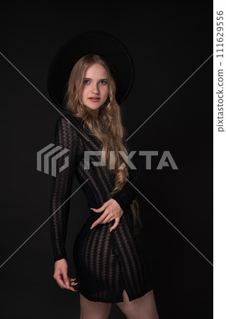 Slim woman striking graceful pose with one hand on waist. Black background accentuates her beauty, while her black short knitted translucent dress and wide brimmed felt hat add touch of elegance Slim woman striking graceful pose with one hand on waist. Black background accentuates her beauty, while her black short knitted translucent dress and wide brimmed felt hat add touch of elegance 111629556