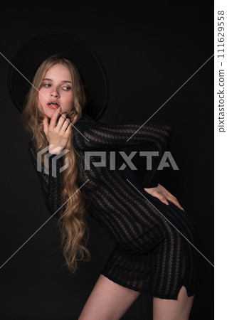 Portrait of young blonde woman striking graceful pose, looking coyly to side with finger to lips, one hand on waist. Model in short black knitted see-through tight dress, felt hat on black background 111629558