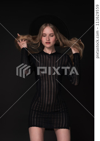 Portrait slim young blonde woman with closed eyes and hands in motion straightening long hair. Model in short tight knitted translucent dress and wide-brimmed felt hat. Black background adds to drama 111629559