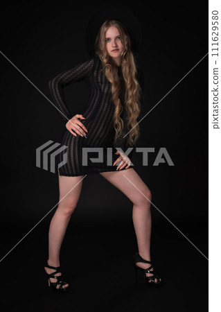 Full length portrait portrait of blonde model exudes confidence as she strikes pose on black background, gazing at camera. Fashionista is rocking chic black dress, complete with wide-brimmed felt hat 111629580