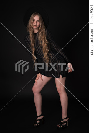 Full length portrait of sexy blonde young woman with long legs on black background. Blonde model with long hair wears black short knitted transparent dress, wide brimmed felt hat, high heels shoes Full length portrait of sexy blonde young woman with long legs on black background. Blonde model with long hair wears black short knitted transparent dress, wide brimmed felt hat, high heels shoes 111629581