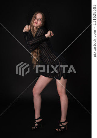 Breathtaking young woman striking confident pose with arms crossed at shoulder height on black background. Blonde with long hair and short knitted black dress, wide-brimmed felt hat, high heeled shoes Breathtaking young woman striking confident pose with arms crossed at shoulder height on black background. Blonde with long hair and short knitted black dress, wide-brimmed felt hat, high heeled shoes 111629583