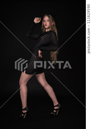 Full length portrait of woman is posing in front of black background. Blonde model with long hair is wearing black short tight knitted transparent dress, wide-brimmed felt hat and high heeled shoes Full length portrait of woman is posing in front of black background. Blonde model with long hair is wearing black short tight knitted transparent dress, wide-brimmed felt hat and high heeled shoes 111629590