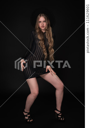 Caucasian woman with long legs, looking gorgeous in black short tight knitted transparent tight dress, wide-brimmed felt hat and high-heeled shoes. Black background really makes her stand out Caucasian woman with long legs, looking gorgeous in black short tight knitted transparent tight dress, wide-brimmed felt hat and high-heeled shoes. Black background really makes her stand out 111629601