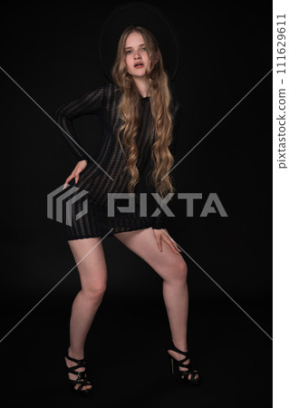 Full length of young woman with long legs, posing on black background. Blonde model with long hair is wearing black short knitted transparent tight dress, wide brimmed felt hat and high heeled shoes Full length of young woman with long legs, posing on black background. Blonde model with long hair is wearing black short knitted transparent tight dress, wide brimmed felt hat and high heeled shoes 111629611