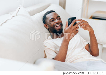 Happy African American man holding a black mobile phone, sitting on a modern sofa in his apartment He is smiling while typing a message and using the internet, enjoying the comfort of his home With a 111631115