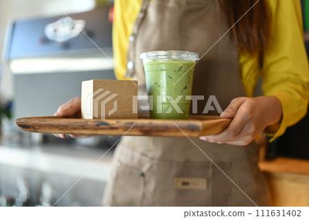 Young woman waiter in apron holding tray with Iced matcha latte to serve customer Young woman waiter in apron holding tray with Iced matcha latte to serve customer 111631402