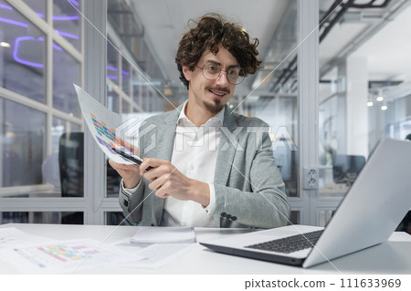 A young, experienced businessman with curly hair attentively examines graphs while working in a bright office environment. A young, experienced businessman with curly hair attentively examines graphs while working in a bright office environment. 111633969