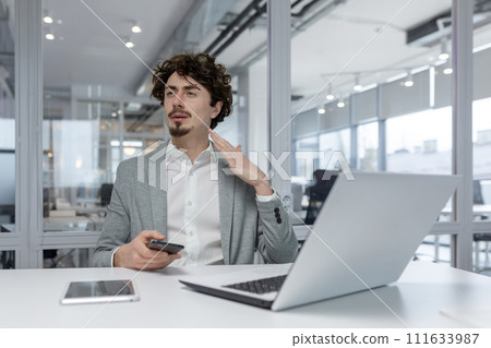Focused young adult businessman with curly hair working at his desk in a modern office environment, using laptop and smartphone. 111633987