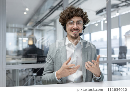 Enthusiastic young businessman with curly hair and headset communicates while working at his office desk. Enthusiastic young businessman with curly hair and headset communicates while working at his office desk. 111634010