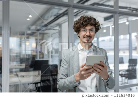 Professional young adult with curly hair, smart business attire, engaged with tablet inside bright office setting. 111634016