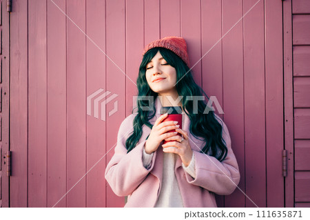 Stylish young smiling hipster woman with closed eyes wearing color hair, pink coat, hat holding reusable coffee cup leaning on beach hut and enjoying moment. Simple pleasures and personal fulfillment Stylish young smiling hipster woman with closed eyes wearing color hair, pink coat, hat holding reusable coffee cup leaning on beach hut and enjoying moment. Simple pleasures and personal fulfillment 111635871